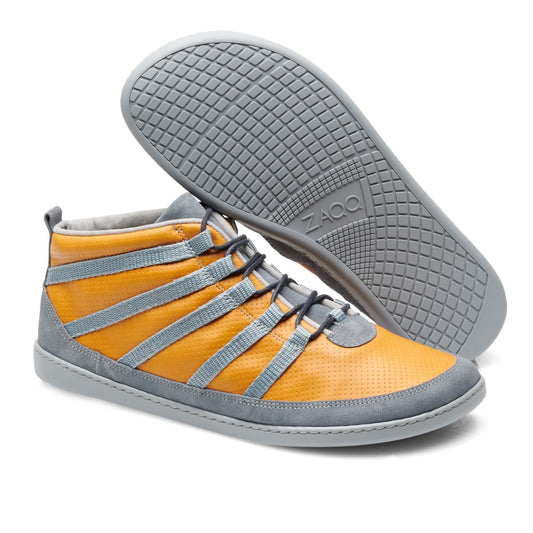 Ein Paar trendige SPARQ Mid Yellow-Sneaker von ZAQQ mit orangefarbenem Nappaleder und grauen Akzenten im Mid-Top-Stil. Diese Schuhe haben graue Schnürsenkel und eine strapazierfähige graue Gummisohle, wobei ein Schuh kunstvoll auf die Seite gelegt wurde, um das komplizierte Muster der Sohle hervorzuheben.