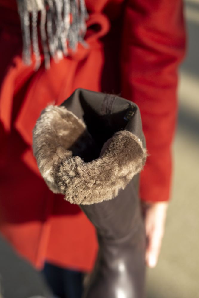 Nahaufnahme einer Person in einem roten Mantel, die den QATCH Winter Brown-Stiefel von ZAQQ hält, der aus luxuriösem Nappaleder gefertigt ist und innen mit kuscheligem Lammfell gefüttert ist. Der Hintergrund ist verschwommen und hebt die exquisite Textur und das weiche Futter dieses Barfuß-Stiefels hervor.