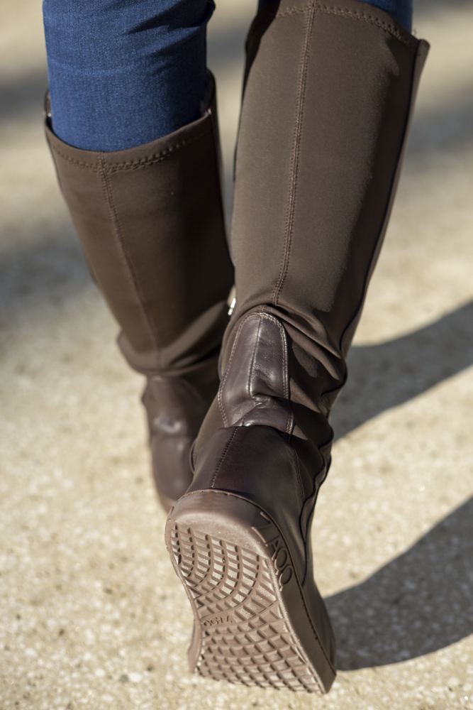 Eine Person trägt kniehohe braune QATCH Winter Brown-Stiefel von ZAQQ und Blue Jeans und läuft auf einem Kiesweg. Das Foto konzentriert sich auf die Stiefel von hinten und hebt das Lammfell-Futter, die strukturierten Sohlen und die sorgfältigen Nähte hervor.