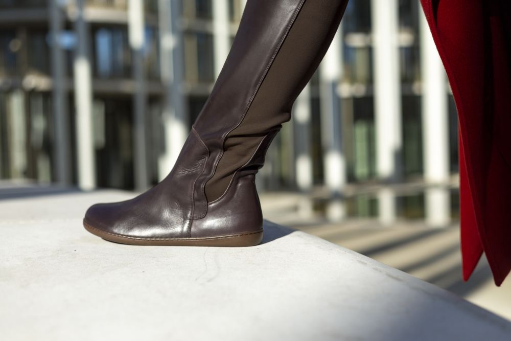 Eine Person in hohen, dunklen ZAQQ QATCH Nappa Brown Lederstiefeln steht auf einem hellen Untergrund. Im Hintergrund sind verschwommene Architekturelemente mit vertikalen Linien zu sehen, seitlich ist ein Teil eines roten Kleidungsstückes zu erkennen.