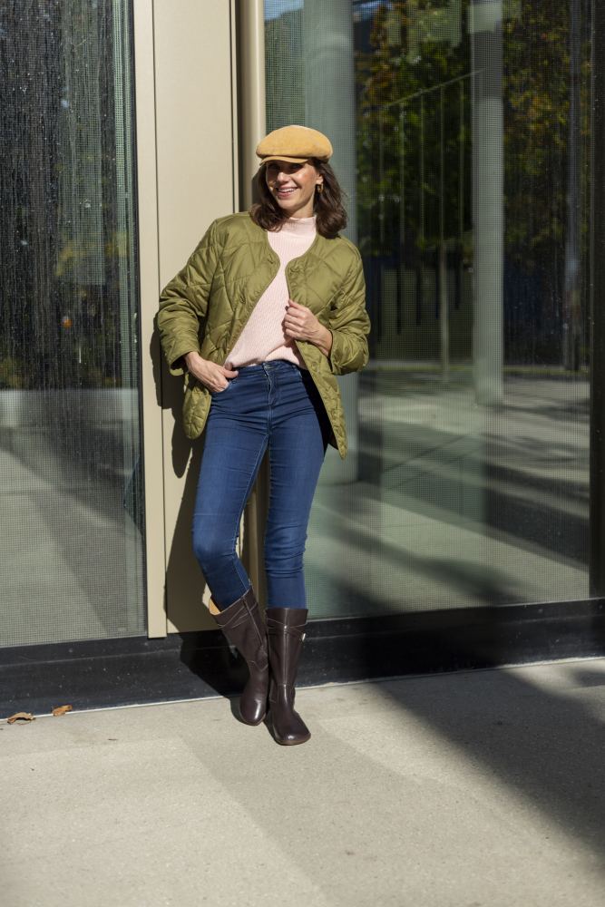 Eine Frau steht im Freien und lehnt an einem modernen Glasgebäude. Sie trägt eine grüne Steppjacke, blaue Jeans und stylische Damenstiefel aus geschmeidigem Ziegenleder, genauer gesagt den ATTRAQT Nappa Brown von ZAQQ. Auf ihrem Kopf sitzt eine beige Schiebermütze, sie lächelt und hält eine Sonnenbrille in der rechten Hand, während das Sonnenlicht sanfte Schatten um sie herum wirft.