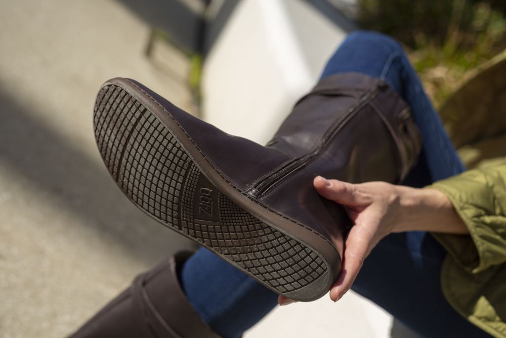 Eine Person in Blue Jeans und grüner Jacke sitzt und hält ihr ausgestrecktes Bein hoch, so dass die Sohle eines ATTRAQT Nappa Brown Damenstiefels von ZAQQ mit Strukturmuster und sichtbarem Markenlogo zu sehen ist. Der Hintergrund ist unscharf.