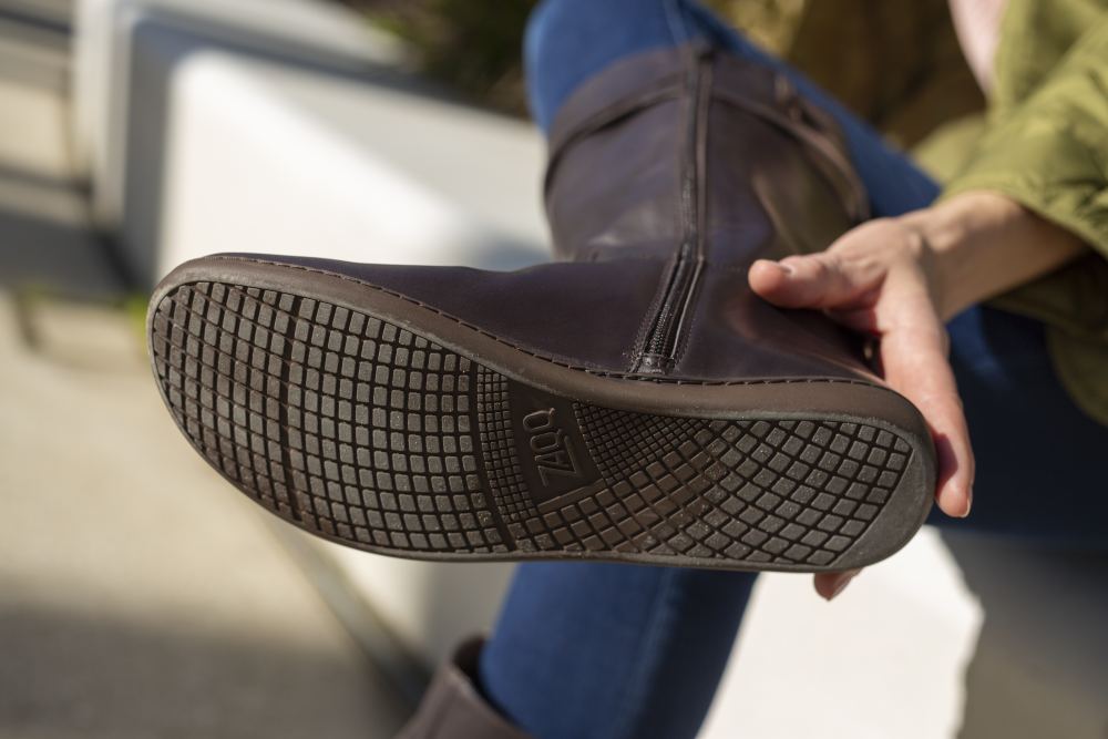 Eine sitzende Person hält ihr Bein hoch und zeigt die Sohle eines ATTRAQT Nappa Brown Damenstiefels mit einem strukturierten Muster mit dem Markennamen ZAQQ. Der braune Stiefel aus hochwertigem Ziegenleder passt perfekt zu ihren blauen Jeans und ihrer grünen Jacke.