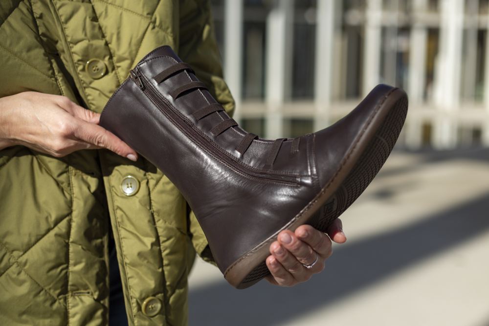 Eine Person in einer grünen Steppjacke hält den ZAQQ ATTRAQT Stretch Nappa Brown aus glattem braunem Leder mit seitlichen Reißverschlüssen und Riemendetails. Der Hintergrund ist sanft verschwommen und suggeriert eine Outdoor-Kulisse.