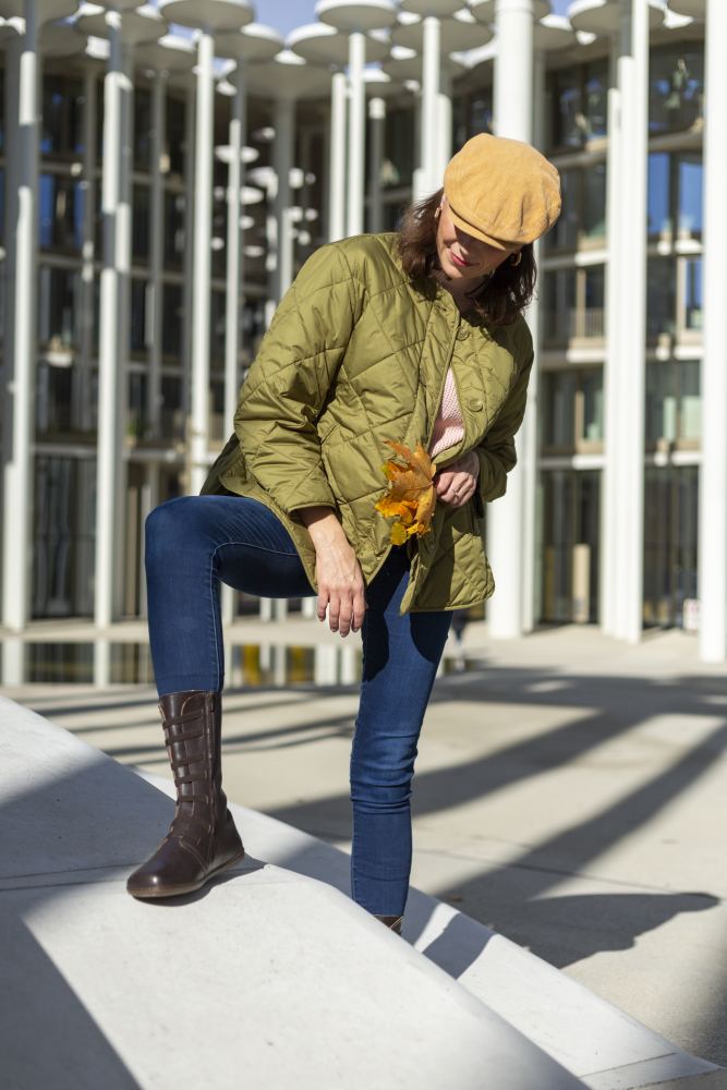 Eine Person in einer grünen Steppjacke, Blue Jeans und robusten ATTRAQT Stretch Nappa Brown-Schuhen von ZAQQ steht auf einer Betonstufe und hält Herbstblätter in den Händen. Sie trägt eine hellbraune Kappe und befindet sich draußen vor einem modernen Gebäude mit Glas und weißen Säulen.