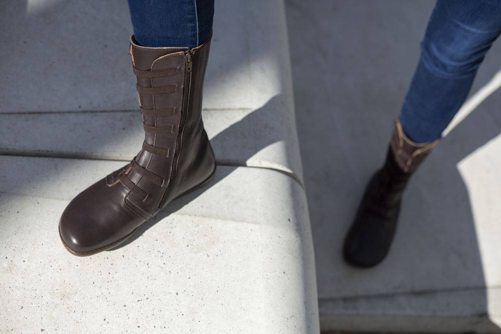 Eine Person trägt die braunen, kniehohen ATTRAQT Stretch Nappa-Stiefel von ZAQQ und Blue Jeans und geht Betonstufen hinauf. Die Stiefel mit flexiblen Riemen und Reißverschlüssen an den Seiten fangen das Sonnenlicht ein, während Schatten über die Stufen tanzen.