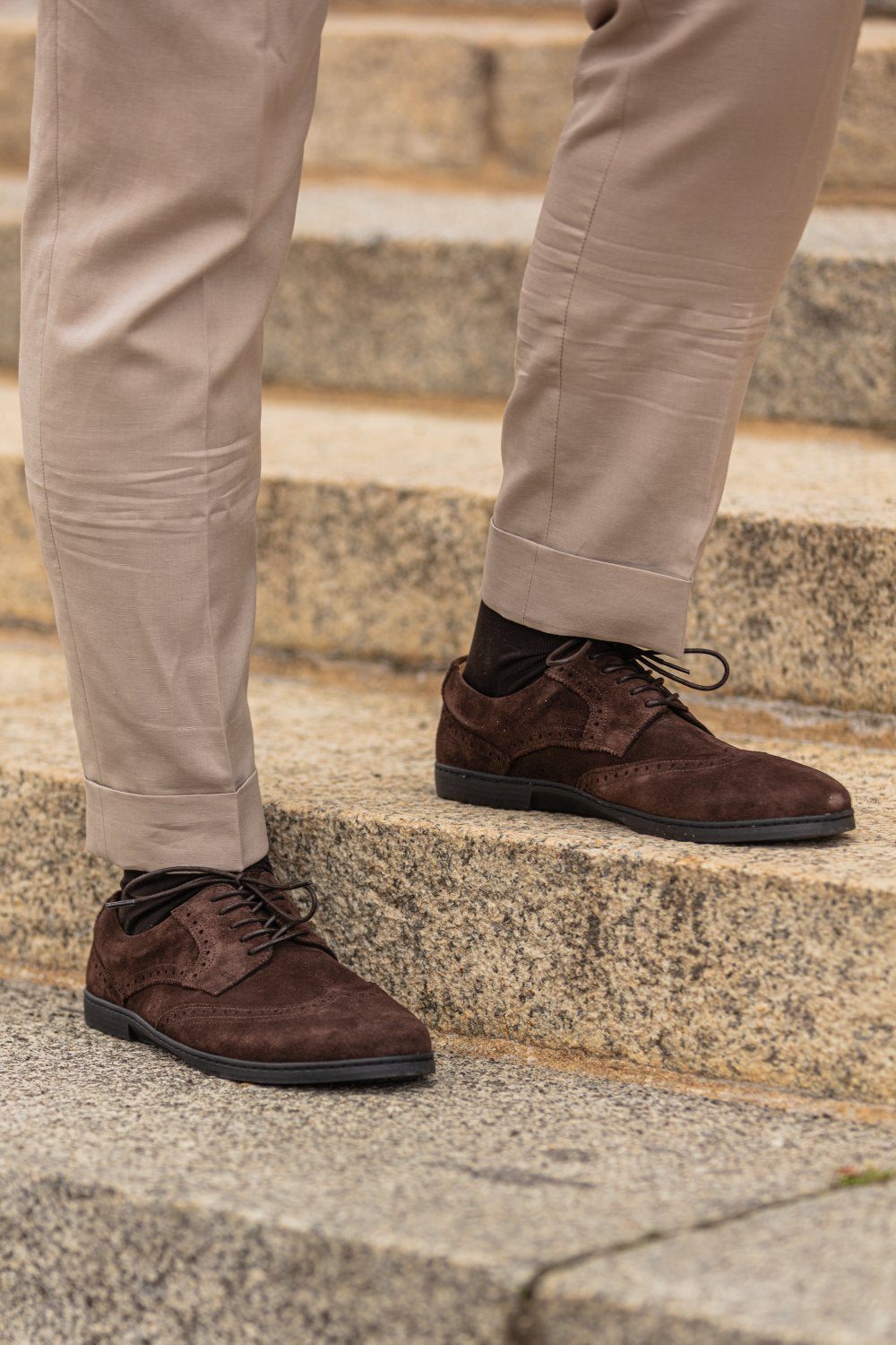 Jemand trägt beige Hosen und dunkelbraune BRIQ Brogue Velours Brown-Abendschuhe von ZAQQ, gefertigt aus französischem Kalbleder, und steht auf Steinstufen. Die Schuhe haben schwarze Schnürsenkel und werden elegant mit dunklen Socken kombiniert.