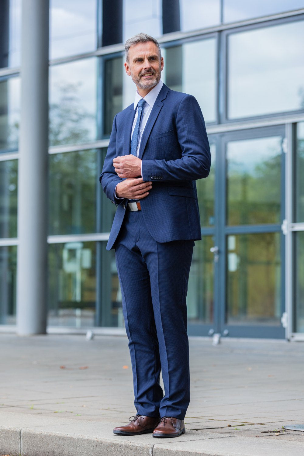 Ein bärtiger Mann im blauen Anzug, der die Schuhe DUQE Antique Brown von ZAQQ aus atmungsaktivem Glattleder trägt, steht vor einem modernen Glasgebäude. Nachdenklich rückt er sein Jackett zurecht.