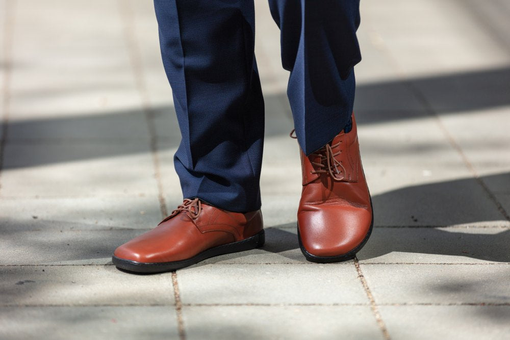 Eine Person in PEAQ Cognac Lederschuhen von ZAQQ und marineblauer Hose läuft auf einem sonnenbeschienenen Steinweg. Die Schuhe, die mit Barfußsohle-Komfort ausgestattet und mit braunen Schnürsenkeln verziert sind, werfen Schatten auf den Boden.