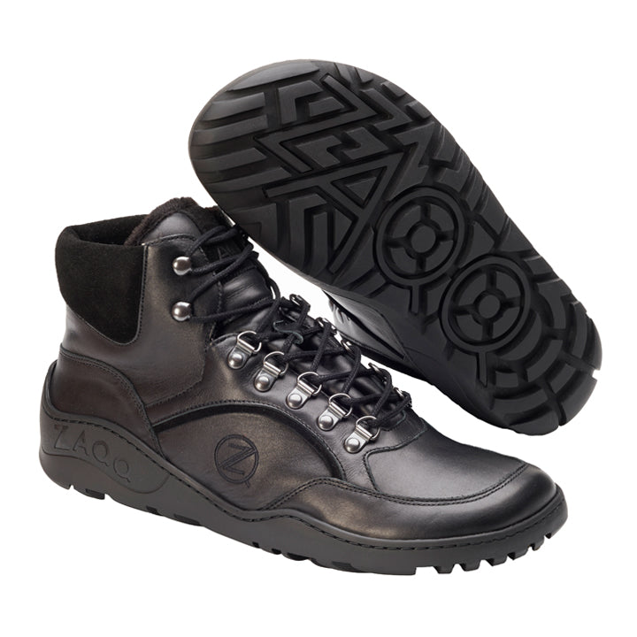 Die TERRAQ Black Winter Waterproof-Stiefel von ZAQQ im Wanderstil aus schwarzem Leder verfügen über verstärkte Knöchel und Metallösen für Schnürsenkel. Sie erinnern an das Design des TERRAQ-Barfußschuhs. Die robuste geometrische Sohle bietet hervorragenden Halt und Wärme für jedes Abenteuer. Ein Stiefel steht aufrecht, während der andere sein charakteristisches Sohlenmuster zur Schau stellt.