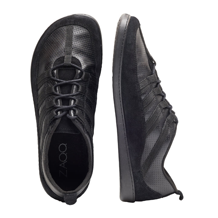 Ein Paar SPARQ Low Black-Sneaker mit perforiertem Design. Ein Schuh ist von oben zu sehen und zeigt die Schnürsenkel und den Markennamen ZAQQ im Inneren, während der andere Sneaker von der Seite betrachtet wird und sein schlankes Profil und seine strukturierte Nappalederoberfläche zur Schau stellt.