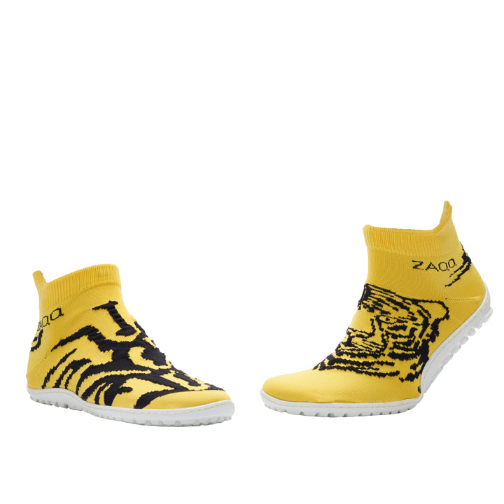 Die ZAQQ SOQQ Tiger Yellow-Schuhe bestechen durch ein auffälliges schwarzes Tigerstreifenmuster auf leuchtend gelben Slippern, akzentuiert durch flexible weiße Sohlen und ein knöchelhohes Design. Der Markenname ZAQQ ist prominent auf der Seite dieser modischen Barfußschuhe angebracht. Sie sind fachmännisch auf Komfort ausgelegt und präsentieren sich elegant vor einem weißen Hintergrund.