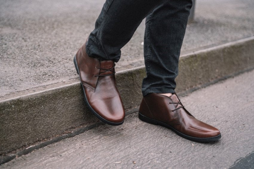 Nahaufnahme einer Person in dunkler Hose und schicken braunen Lederstiefeletten, die mit einem Fuß auf dem Bordstein und dem anderen auf der Straße steht. Die Stiefel sind elegant mit Schnürsenkeln und bilden einen Kontrast zum strukturierten Bürgersteig, was eine Mischung aus urbanem Stil und Eleganz suggeriert.