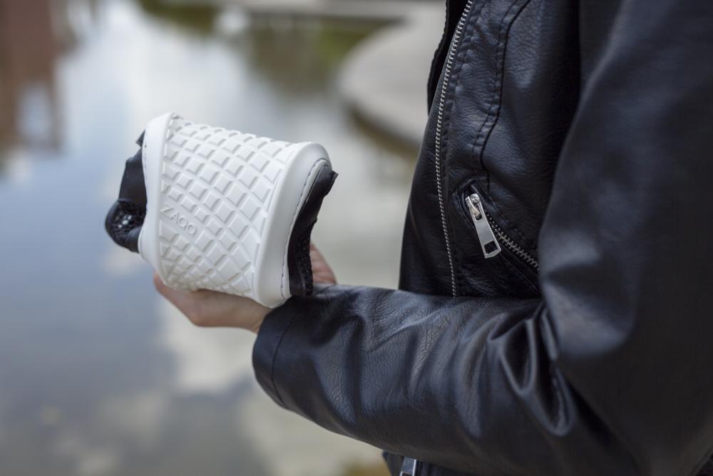 Eine Person in einer schwarzen ZAQQ ALIQANTE Jacke hält einen flexiblen weißen Barfußschuh mit einem Strukturmuster an einem ruhigen Gewässer.