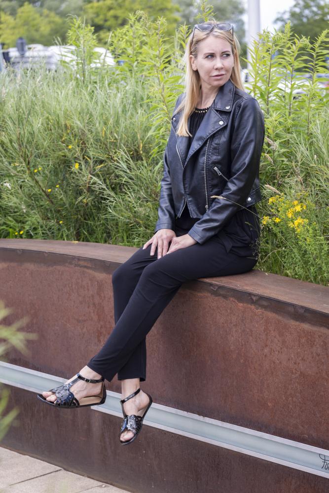 Eine Frau in einer schwarzen BOUQET Jacke und Hose von ZAQQ sitzt auf einer rostigen Metallbank im Freien. Sie trägt eine Sonnenbrille auf dem Kopf und stylische schwarze Barfußsandale, im Hintergrund grüne Büsche.