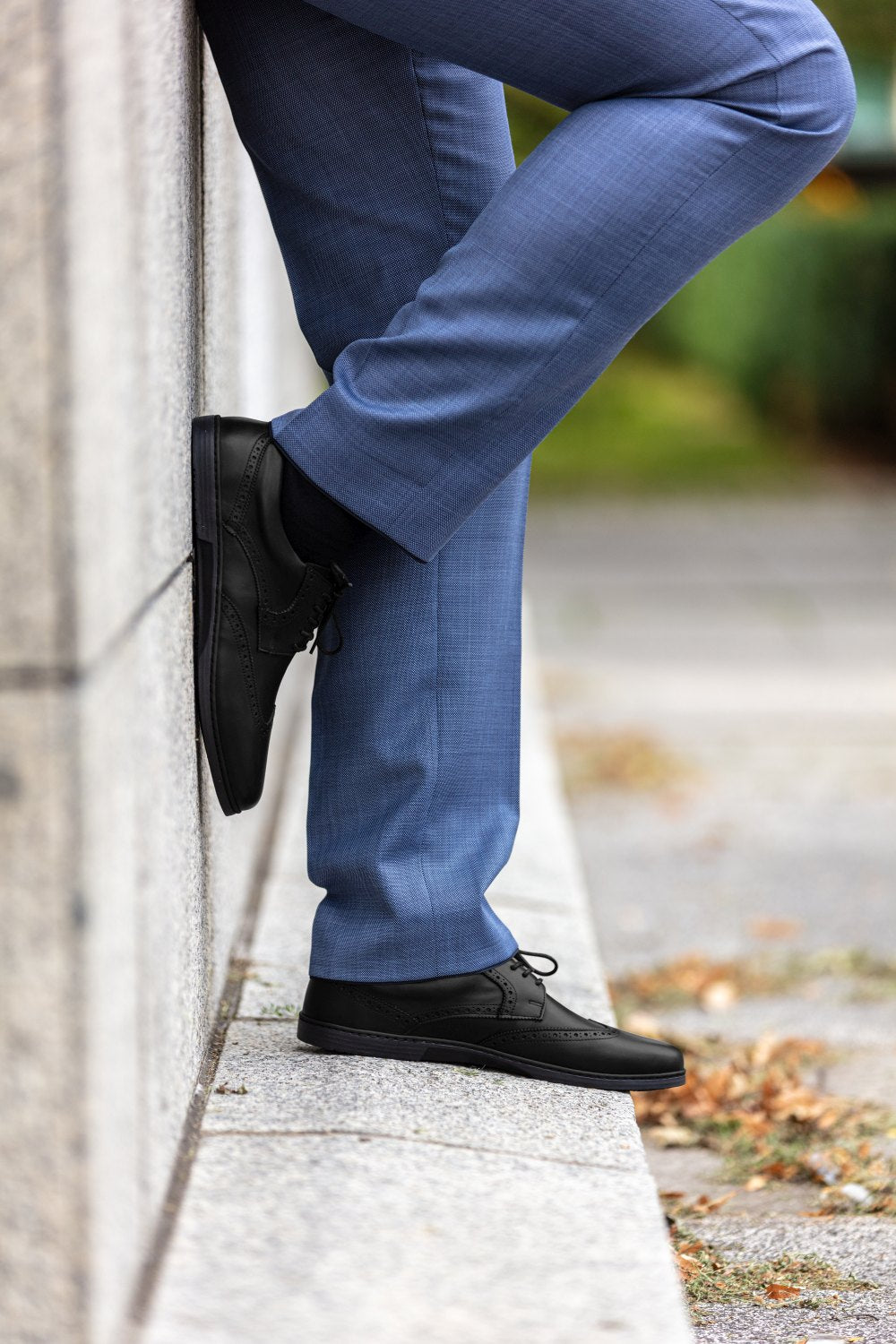 Eine Person in blauen Anzughosen und den klassischen BRIQ Brogue Black-Schuhen von ZAQQ lehnt an einer Steinmauer und steht auf einem Gehweg. Der Hintergrund ist leicht verschwommen und zeigt Grünpflanzen und gefallene Blätter.