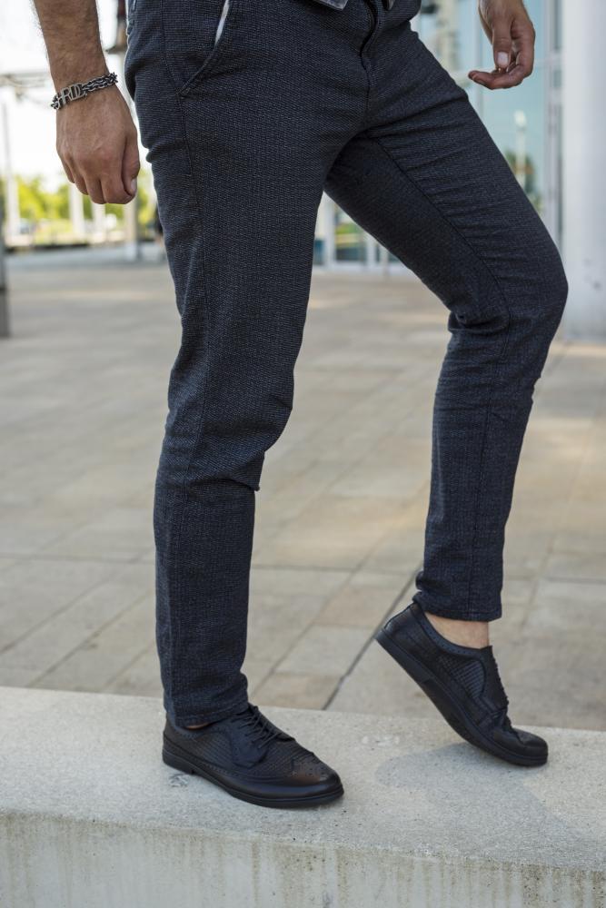 Eine Person in karierten Hosen und dem Schuhwerk BRIQ Brogue Tresse Black von ZAQQ steht auf einem Felsvorsprung vor einer urbanen Kulisse und präsentiert einen stilvollen Business-Casual-Look. Ihr linkes Bein ist angewinkelt, während ihr rechtes Bein gerade auf dem Boden bleibt.