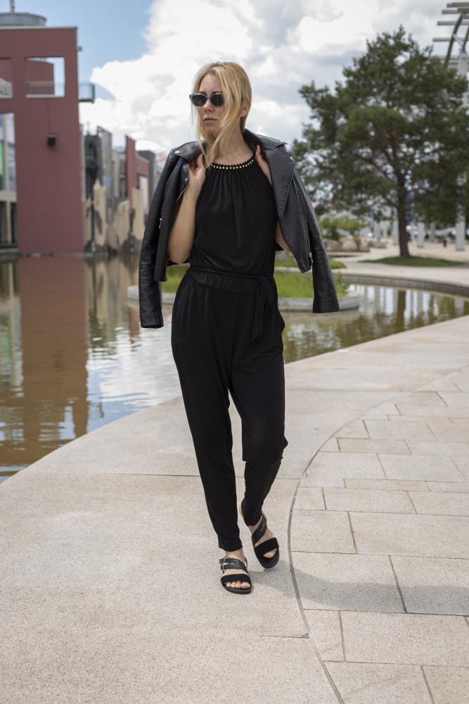 Eine Person mit der Sonnenbrille BROOQE Black von ZAQQ und einem stylischen schwarzen Outfit hängt sich eine Jacke aus Nappaleder über die Schultern und läuft einen Weg neben einem reflektierenden Gewässer entlang. Der Himmel ist teilweise bewölkt, im Hintergrund sind moderne Gebäude und Bäume zu sehen.