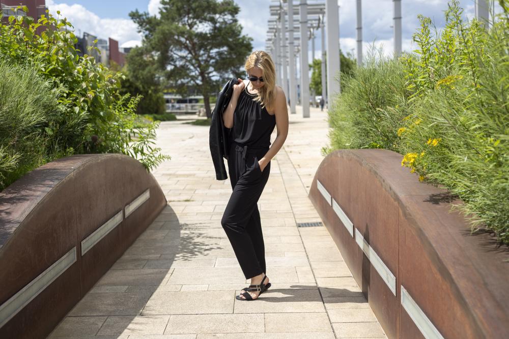 Eine Person im ZAQQ BROOQE Black Outfit läuft selbstbewusst einen von Grün und moderner Architektur gesäumten Gehweg entlang. Stilvoll trägt sie eine Sonnenbrille, eine Jacke über der Schulter und feminine Sandalen aus Nappa- und Veloursleder führen sie durch die helle Szene unter leicht bewölktem Himmel.