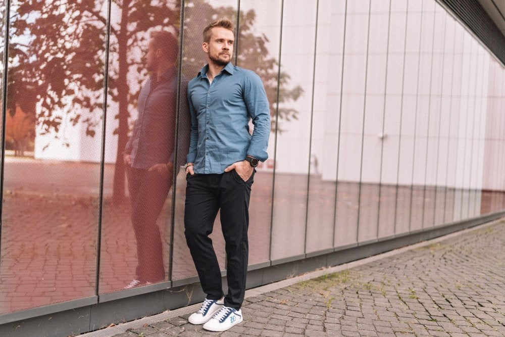 Ein Mann lehnt im Freien an einer reflektierenden Wand. Er trägt ein blaues Hemd mit Knöpfen und eine schwarze Hose und trägt die CEDRIQ Blue Sneakers von ZAQQ. Der Bürgersteig unter ihm ist Kopfsteinpflaster und im Hintergrund sind schwach Herbstbäume zu erkennen.