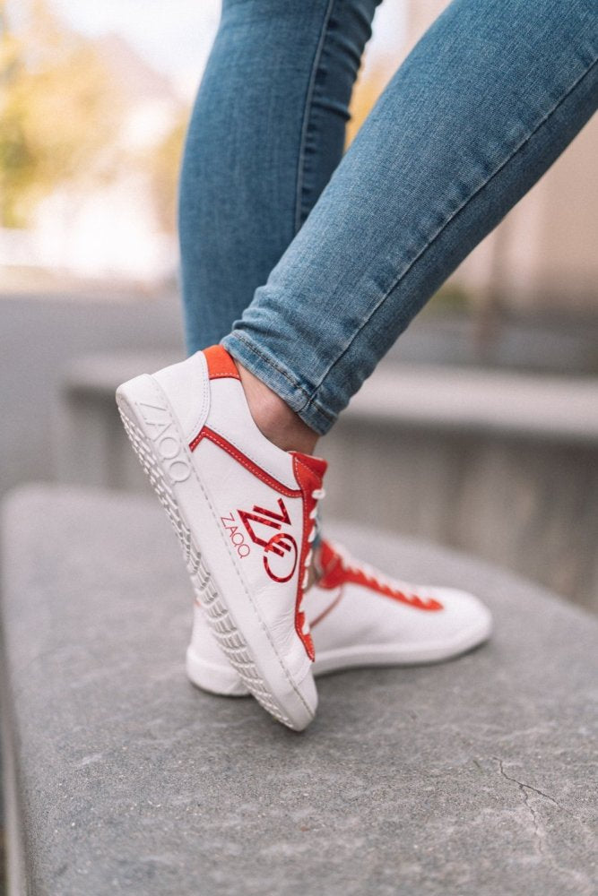 Eine Person in Blue Jeans lehnt auf einer Steinfläche und trägt die CEDRIQ Red Sneakers von ZAQQ, die auffällige rote Akzente und ein sichtbares Logo aufweisen. Die Sneakers verfügen über eine Kautschuksohle für optimalen Komfort. Der unscharfe Hintergrund betont eine Outdoor-Kulisse.