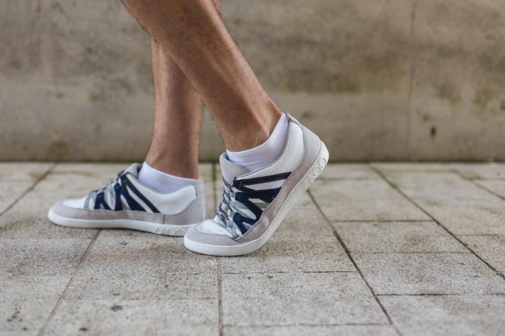 Eine Person in den schicken Sneakern DRAQE White Grey von ZAQQ aus hochwertigem Nappaleder schlendert über einen gefliesten Gehsteig. Die Betonung liegt auf Beinen und Schuhen, im Hintergrund eine Betonwand.