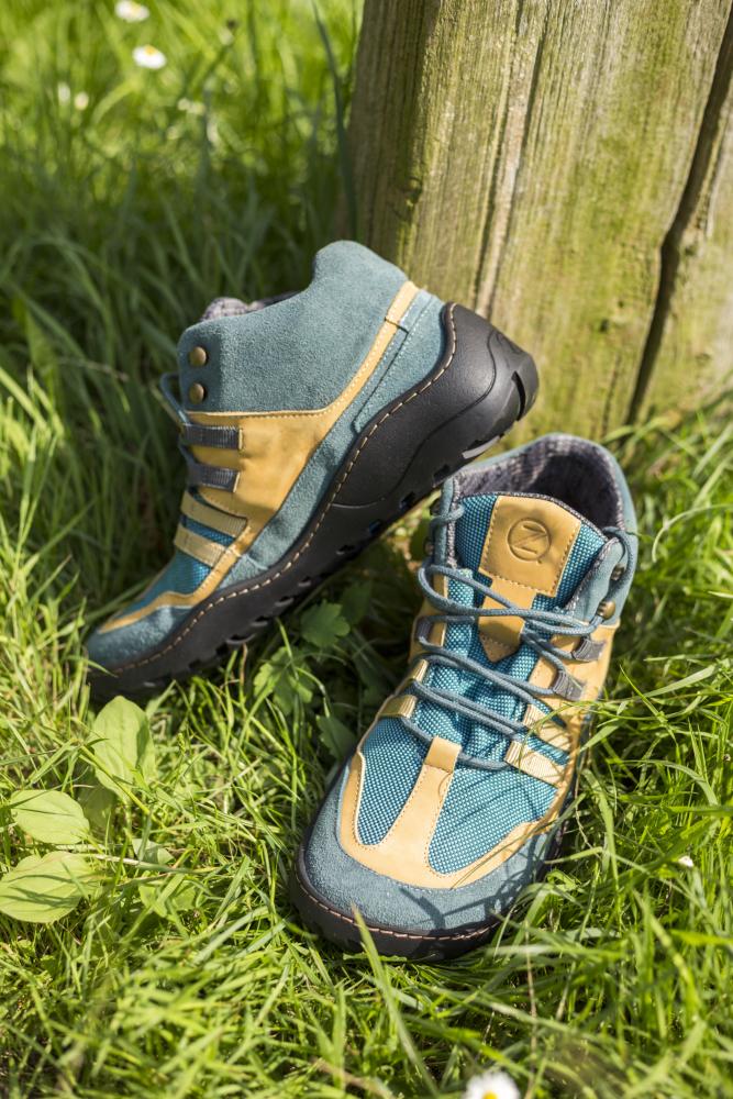 Ein Paar blau-orangefarbene wasserdichte ESQAPE-Wanderschuhe mit den markanten schwarzen Sohlen von ZAQQ stehen auf grünem Gras neben einem Holzpfosten. Ein Schuh steht aufrecht, während der andere lässig am Pfosten lehnt. Kleine weiße Blumen säumen den Bereich und verleihen der Szene eine bezaubernde natürliche Note.