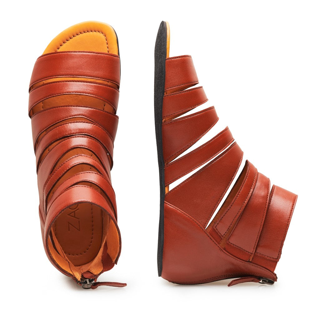 Ein Paar GLADIQ Cognac-Gladiatorsandalen von ZAQQ mit mehreren horizontalen Riemen und Reißverschlüssen auf der Rückseite. Eine Sandale steht aufrecht, während die andere auf der Seite liegt und die elegante flache schwarze Sohle dieser stilvollen Schuhe zur Schau stellt.