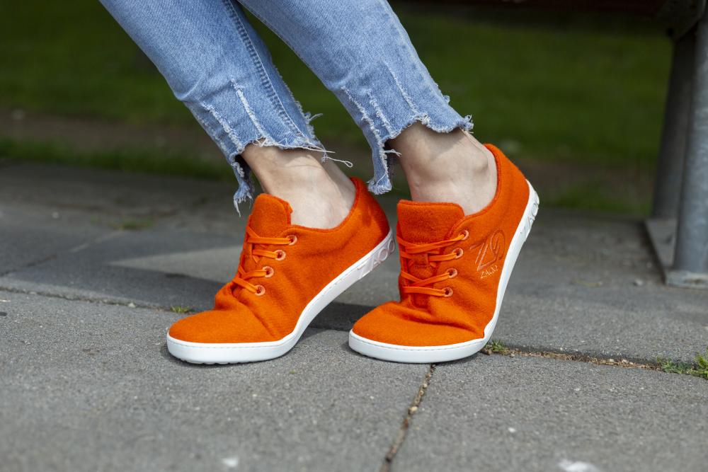 Eine Person, die die LIQE Orange Sneakers von ZAQQ und zerrissene Bluejeans trägt, steht auf einem Steinpflaster und genießt das Barfußgefühl. Das saftige grüne Gras im Hintergrund erinnert an eine parkähnliche Umgebung, in der die natürliche Temperaturregulierung für mehr Komfort sorgt.