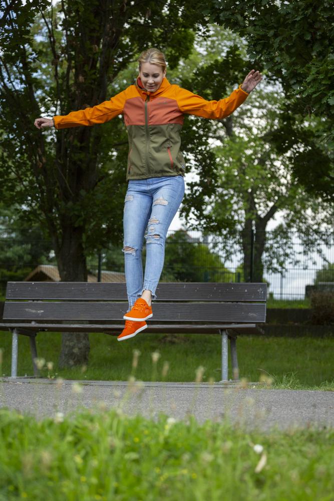 In der leuchtend orangefarbenen Jacke LIQE und den Schuhen von ZAQQ springt eine Person freudig von einer Parkbank und erlebt ein Barfußgefühl. Sie befindet sich im Freien inmitten üppiger grüner Bäume und Gras, und hinter ihnen erstreckt sich der Himmel.