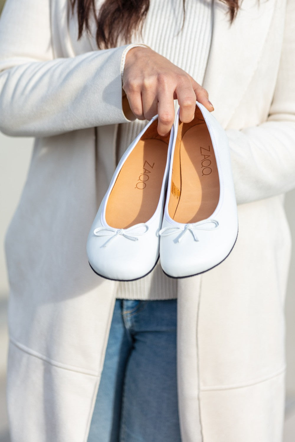 Eine Person im weißen Kittel hält ein Paar weiße Ballerinas von LOOQ, die mit kleinen Schleifen verziert sind. Diese eleganten Ballerinas haben eine Innenausstattung aus beigefarbenem Nappaleder mit dem aufgedruckten Markennamen ZAQQ. Der sanft verschwommene Hintergrund betont die raffinierte Schlichtheit der Schuhe.