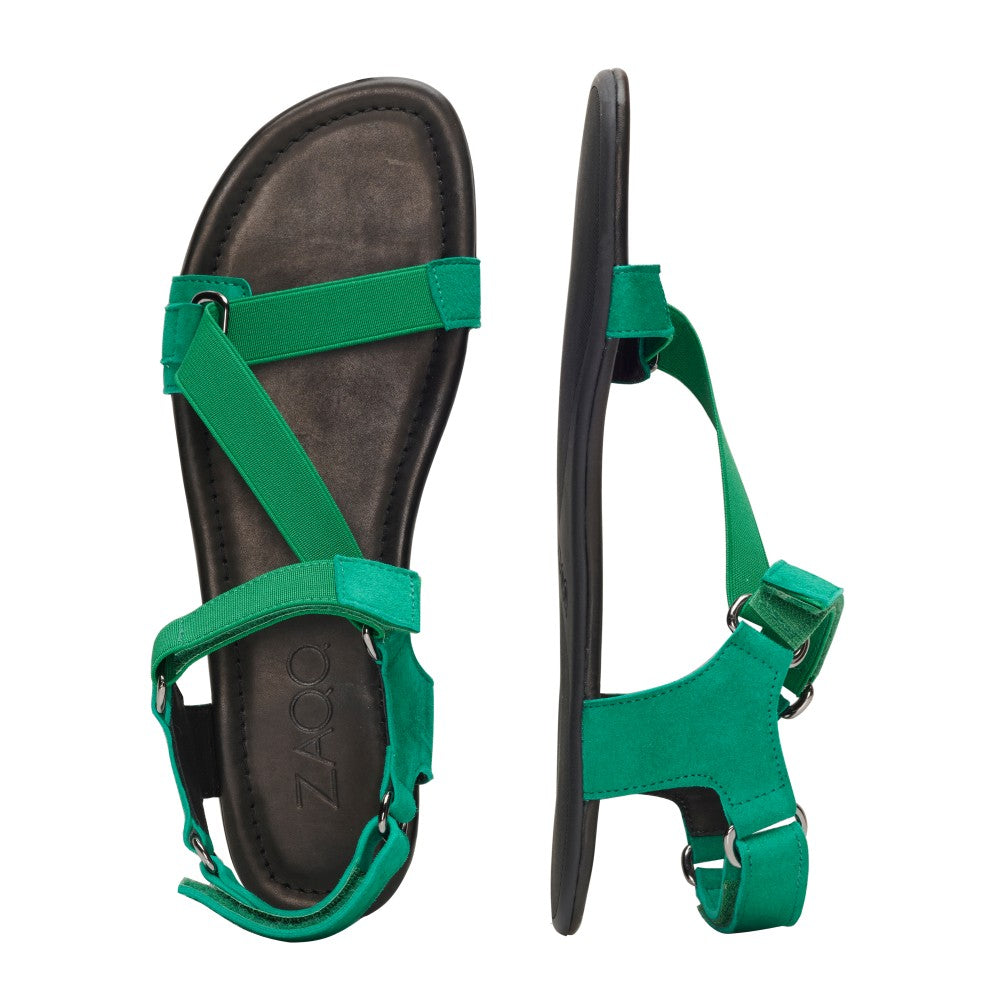 Entdecken Sie die ZAQQ MIQA Green Sandalen aus weichem Material. Eine Sandale liegt flach und zeigt überkreuzte Riemen und eine bequeme Barfußsandale-Sohle, während die andere aufrecht steht und ihr verstellbares Riemchensystem-Fersenband für eine tadellose Passform betont.