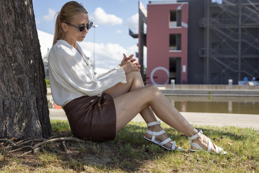 Eine Frau mit Sonnenbrille sitzt im Gras unter einem Baum und strahlt in ihrer weißen Bluse und den braunen Ledershorts eine entspannte Stimmung aus. Ihre weißen ZAQQ MONIQ Barfußsandale passen perfekt zu dem sonnigen Tag, mit einem modernen Gebäude und schimmerndem Wasser als Kulisse.