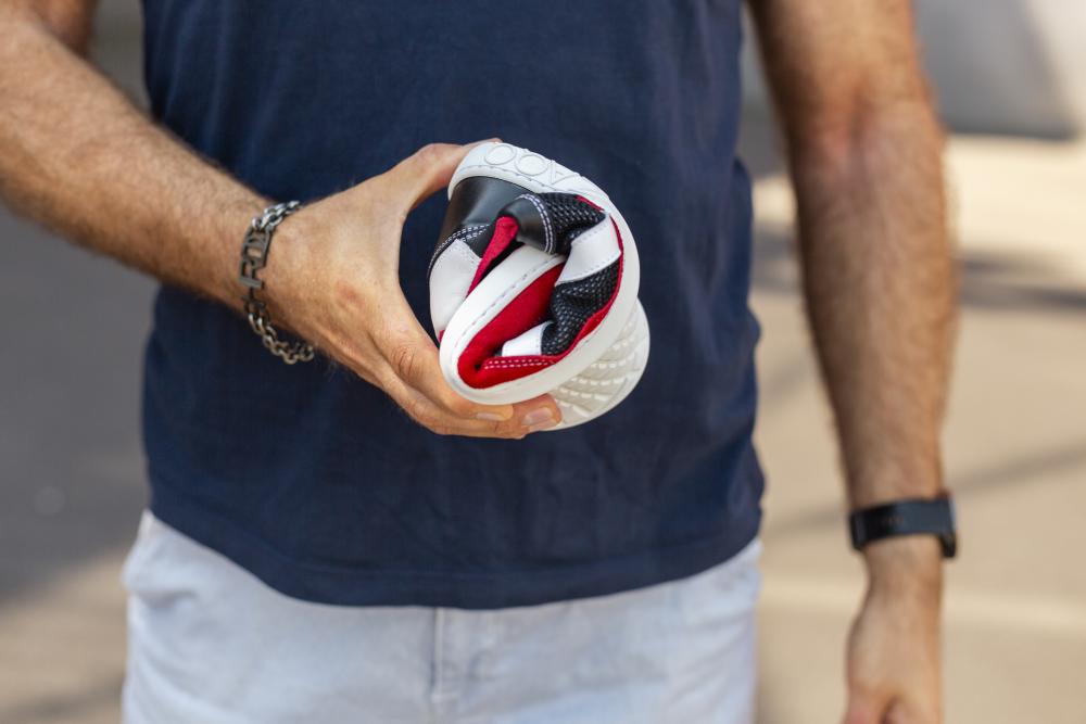 Eine Person in einem marineblauen Hemd und hellen Hosen hält einen flexiblen, aufgerollten OAQLEY Red Barfußschuh mit dem unverwechselbaren rot-weiß-schwarzen Design von ZAQQ. Die Person ist mit einem Armband und einer Uhr geschmückt und genießt den sonnigen Tag im Freien.