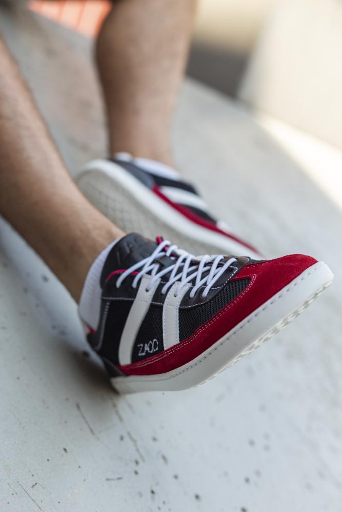 Jemand sitzt draußen auf einer Betonfläche und trägt auffällige ZAQQ-Sneaker mit einem leuchtenden rot-schwarz-weißen Design. Die makellosen weißen Schnürsenkel des Modells OAQLEY Red fallen auf und erinnern an ihren beliebten Stil. Der wunderschön verschwommene Hintergrund verstärkt das elegante Erscheinungsbild dieser auffälligen Sneaker.