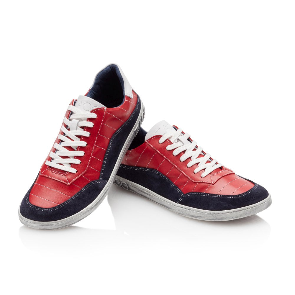 Wir präsentieren den QAMPION Navy Red von ZAQQ, ein Paar stylische Sneaker aus luxuriösem Nappaleder. Diese Sneaker zeichnen sich durch ein auffälliges rotes Obermaterial, weiße Schnürsenkel und marineblaue Akzente an Zehen und Sohlen aus. Kunstvoll auf einem weißen Hintergrund positioniert, lehnt sich ein Sneaker leicht an den anderen.