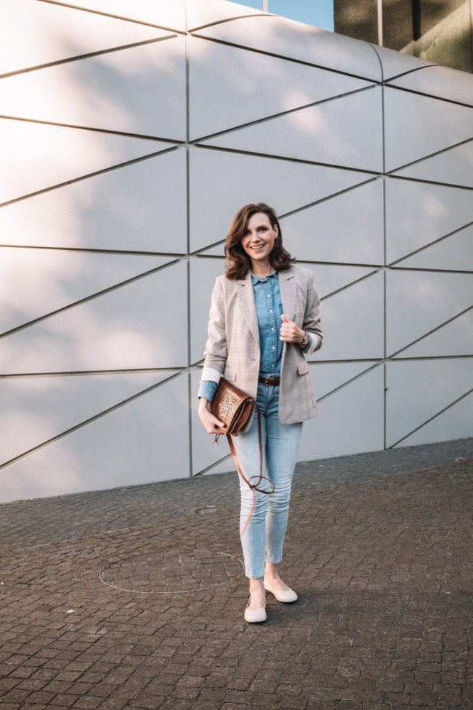 Eine Frau steht vor einer modernen, geometrischen Wand. Sie trägt einen hellkarierten Blazer über einem Jeanshemd, schmale Jeans und schicke weiße QARMA Beige Sneakers von ZAQQ. Sie hält eine braune Handtasche und lächelt in die Kamera.