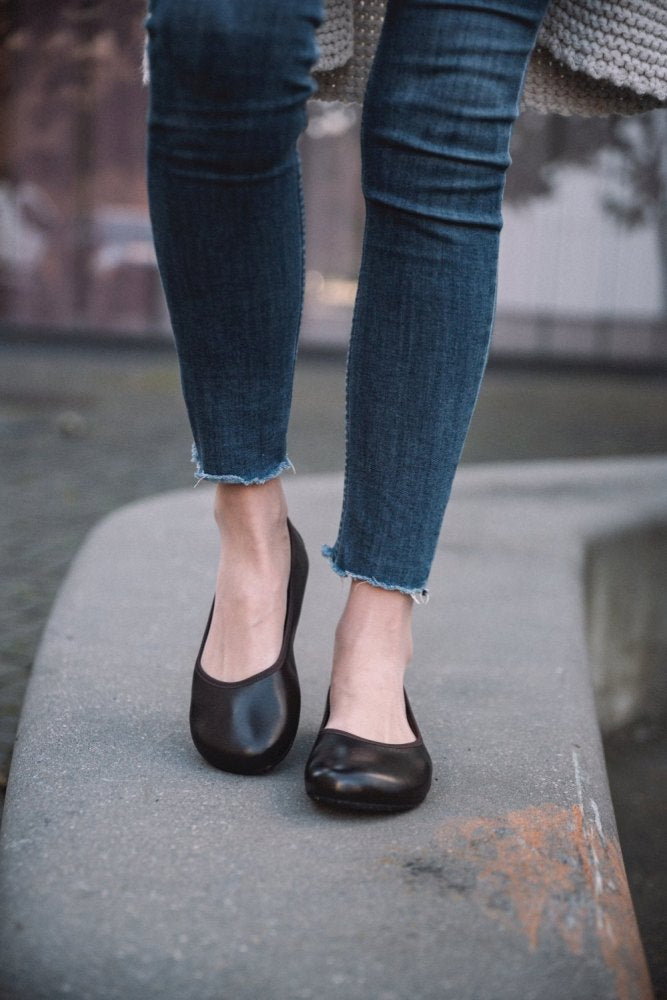 Eine Person in Jeans mit ausgefranstem Saum und stylischen dunkelbraunen ZAQQ QARMA-Ballerinas läuft auf einem Betonvorsprung entlang. Der Hintergrund ist verschwommen und konzentriert sich auf die Füße und Schuhe aus glattem Nappaleder, was einen schicken Barfuß-Ballerina-Look erzeugt.