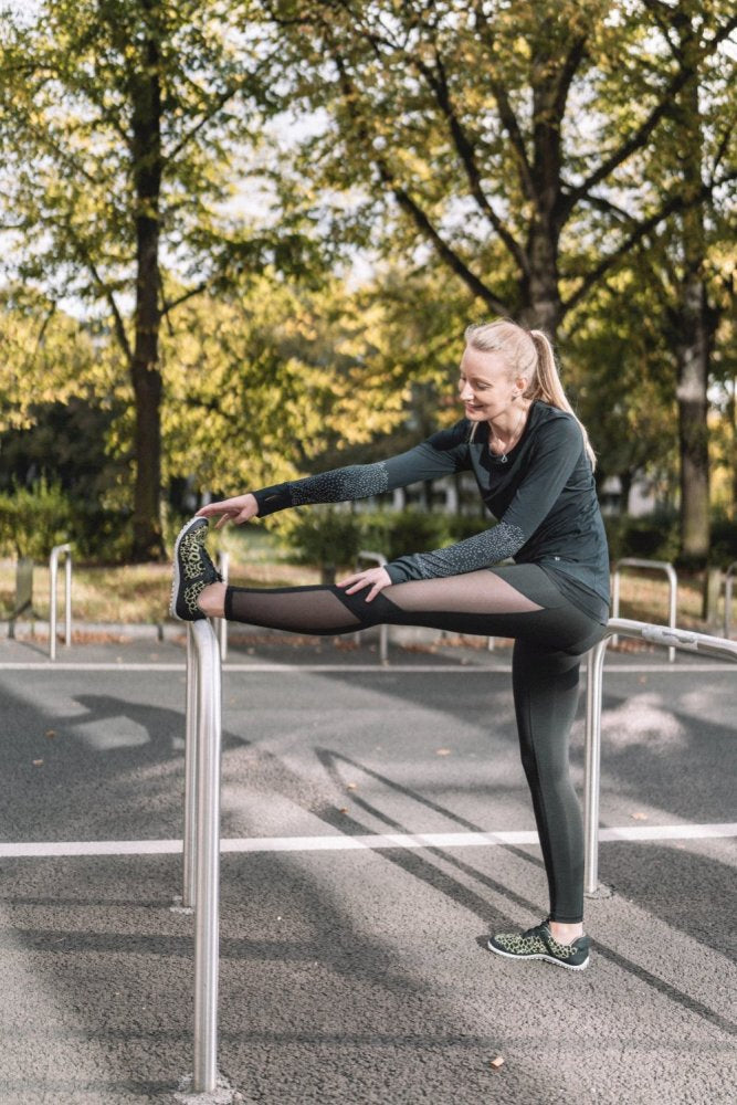 Eine Person in Sportkleidung streckt ihr Bein auf einem Metallgeländer in einem Park im Freien aus. Der sonnendurchflutete Weg hebt ihre QASEY Green-Schuhe von ZAQQ hervor, die aus atmungsaktivem Netzgewebe bestehen. Bäume mit grünen Blättern füllen den Hintergrund und die Sonne wirft Schatten auf den Bürgersteig.