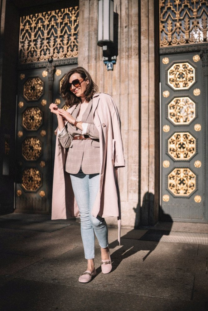 Eine Frau mit Sonnenbrille steht vor einer verzierten Tür mit goldenen Akzenten. Sie trägt einen beigen Trenchcoat, einen karierten Blazer und hellblaue Jeans. Mit einem Lächeln im Gesicht hält sie an diesem sonnigen Tag etwas in der Hand und vervollständigt ihren schicken Look mit den QERP Candy-Schuhen von ZAQQ mit einer bequemen Kautschuksohle aus Veloursleder.