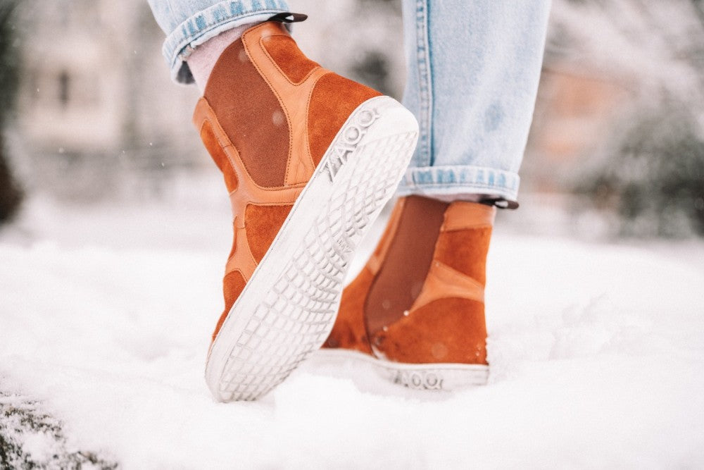 Nahaufnahme einer Person, die ZAQQs QEY WEST Cognac High-Top-Sneakers trägt und im Schnee steht. Die cognacfarbenen Schuhe heben sich vom weißen Schnee ab und die Person trägt hellblaue Jeans, die an den Knöcheln hochgekrempelt sind. Schneeflocken fallen sanft um die Szene herum und verleihen ihr einen Hauch von Winterzauber.