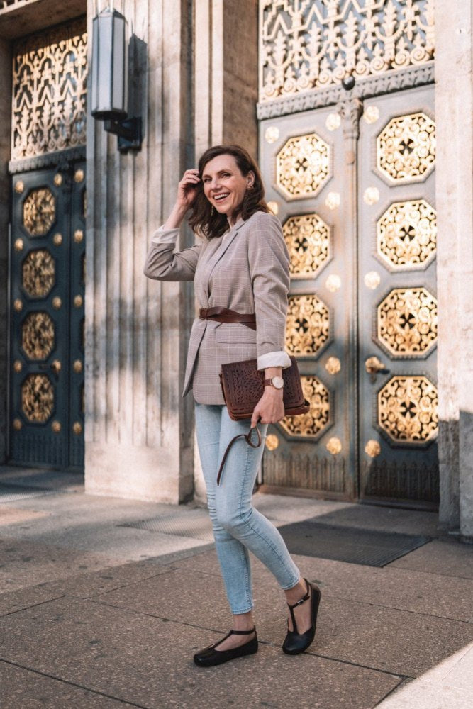 Eine Person in kariertem Blazer und Jeans steht auf der Straße und hält eine strukturierte Clutch in der Hand. Im Hintergrund sind Türen mit aufwendigem Goldmuster zu sehen. Sie trägt eine Uhr am Handgelenk und schicke schwarze Schuhe aus Glattleder von ZAQQ, bekannt als Modell QITE Dark Brown.