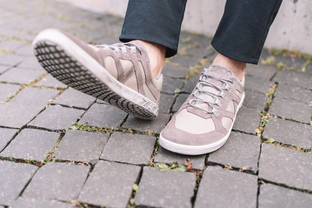 Nahaufnahme einer Person, die die stylischen QUSQO Taupe-Sneaker von ZAQQ trägt. Die gemusterten Sohlen sind aus atmungsaktivem Mesh-Gewebe gefertigt und bieten ein Barfußschuh-Gefühl. Die Person trägt dunkle Hosen vor einer grauen, strukturierten Wand, was die Bewegungsfreiheit auf dem Steinpflaster betont.