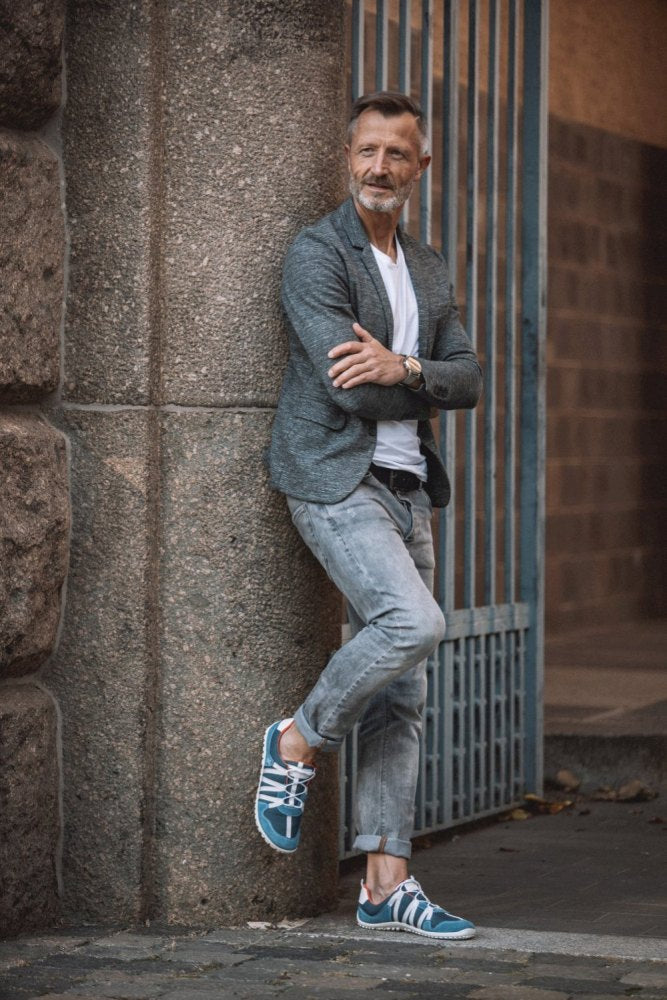 Ein modischer Mann lehnt an einer Steinmauer. Er trägt einen grauen Blazer, ein weißes Hemd und helle Jeans. Seine sportlichen RIQ Blue-Sneaker von ZAQQ sind aus atmungsaktivem Meshgewebe gefertigt. Mit verschränkten Armen und Beinen strahlt er neben einem Eisentor eine lässige und doch stilvolle Ausstrahlung aus.