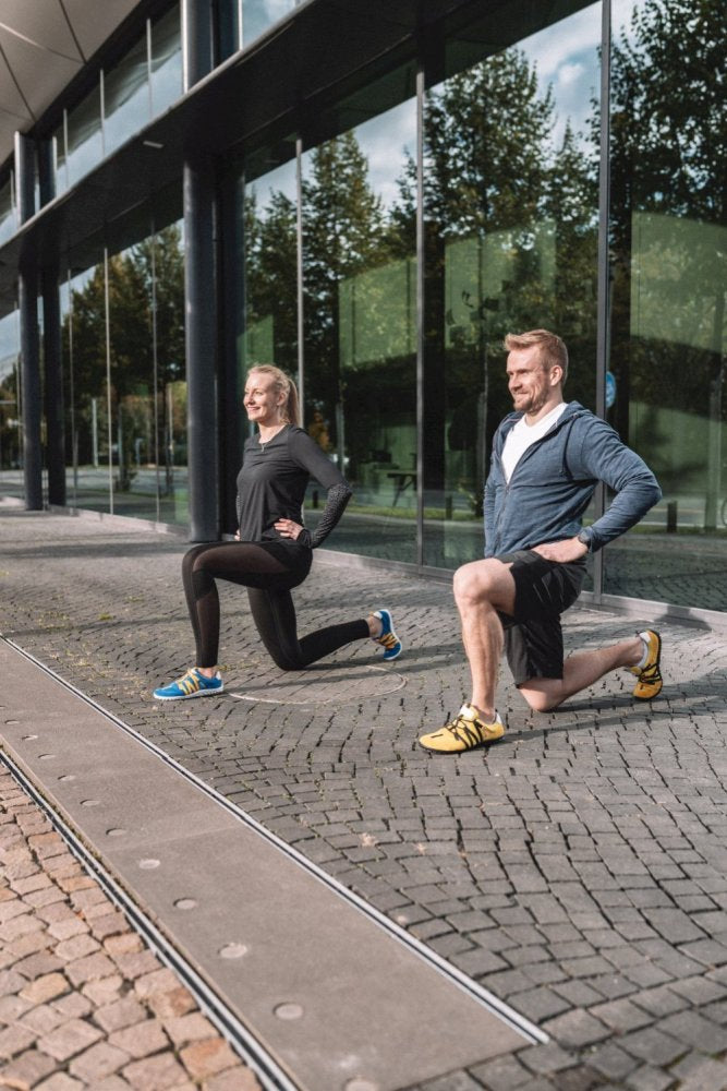 Zwei Personen in Sportbekleidung, mit Akzenten in RIQ Yellow von ZAQQ, machen Ausfallschritte auf einem Kopfsteinpflaster neben einem modernen Glasgebäude. Sie genießen den flexiblen Komfort ihrer Barfußschuhe und trainieren gerne an diesem sonnigen Tag im Freien, während sich die Bäume wunderschön in den Fenstern spiegeln.