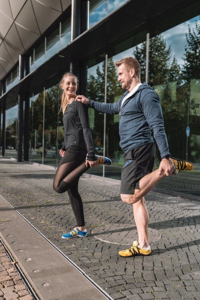 Ein Mann und eine Frau strecken sich draußen auf einem gepflasterten Gehweg neben einem modernen Glasgebäude. In eleganter Sportkleidung und RIQ Yellow-Schuhen von ZAQQ streckt die Frau ihr rechtes Bein, während der Mann sich auf sein linkes konzentriert. Bäume spiegeln sich in den Fenstern und unterstreichen die ruhige Umgebung.