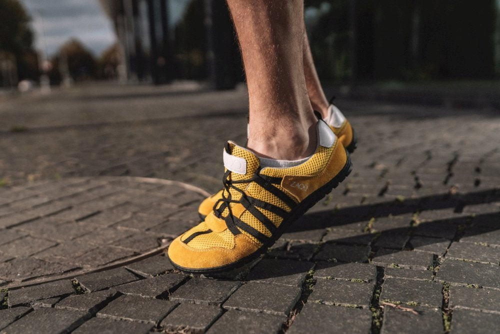 Nahaufnahme einer Person, die RIQ Yellow-Laufschuhe von ZAQQ mit eleganten schwarzen Schnürsenkeln trägt und auf einem Kopfsteinpflasterweg läuft. Diese Barfußschuhe bieten ein flexibles Tragegefühl. Der Hintergrund ist leicht verschwommen und zeigt Bäume und Gebäude in der Ferne.