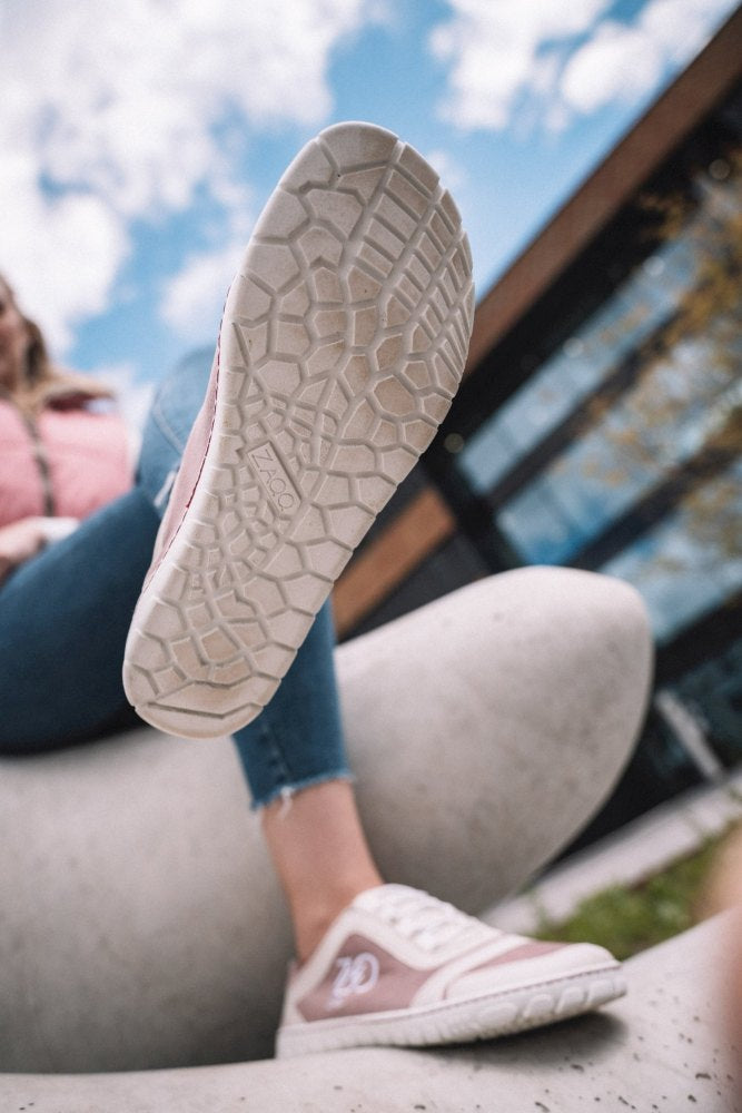 Eine Person präsentiert den ZAQQ SQILL Rose Sneaker mit strukturierter Sohle im Freien. Der Fokus liegt auf dem erhobenen Fuß vor einem Hintergrund aus blauem Himmel, Wolken und moderner Architektur. Dieser rosa-weiße Sneaker aus veganer Mikrofaser passt perfekt zu stylischen Jeans.