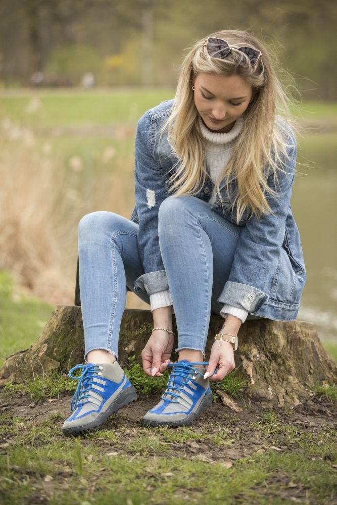 Eine Person sitzt auf einem Baumstumpf in einem Park und bindet die Schnürsenkel ihrer SQOUT Grey Blue Waterproof Sneakers von ZAQQ. Sie trägt eine Jeansjacke und Jeans und trägt eine Sonnenbrille auf dem Kopf. Im Hintergrund sind Gras, Wasser und verschwommene Bäume zu sehen.