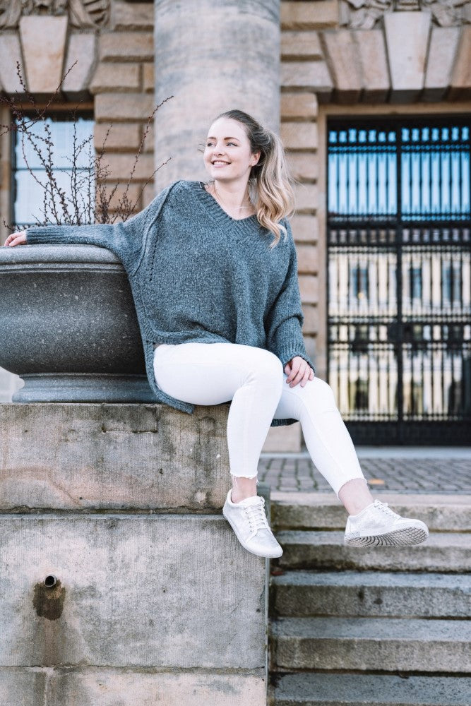 Eine Frau in einem grauen Pullover und weißen Hosen, die stylischen TIQQ Silver White Sneakers von ZAQQ trägt, sitzt mit überkreuzten Beinen auf einem Steinvorsprung und lächelt. Sie steht im Freien vor einem Gebäude mit großen Säulen und einem verzierten Eisentor.